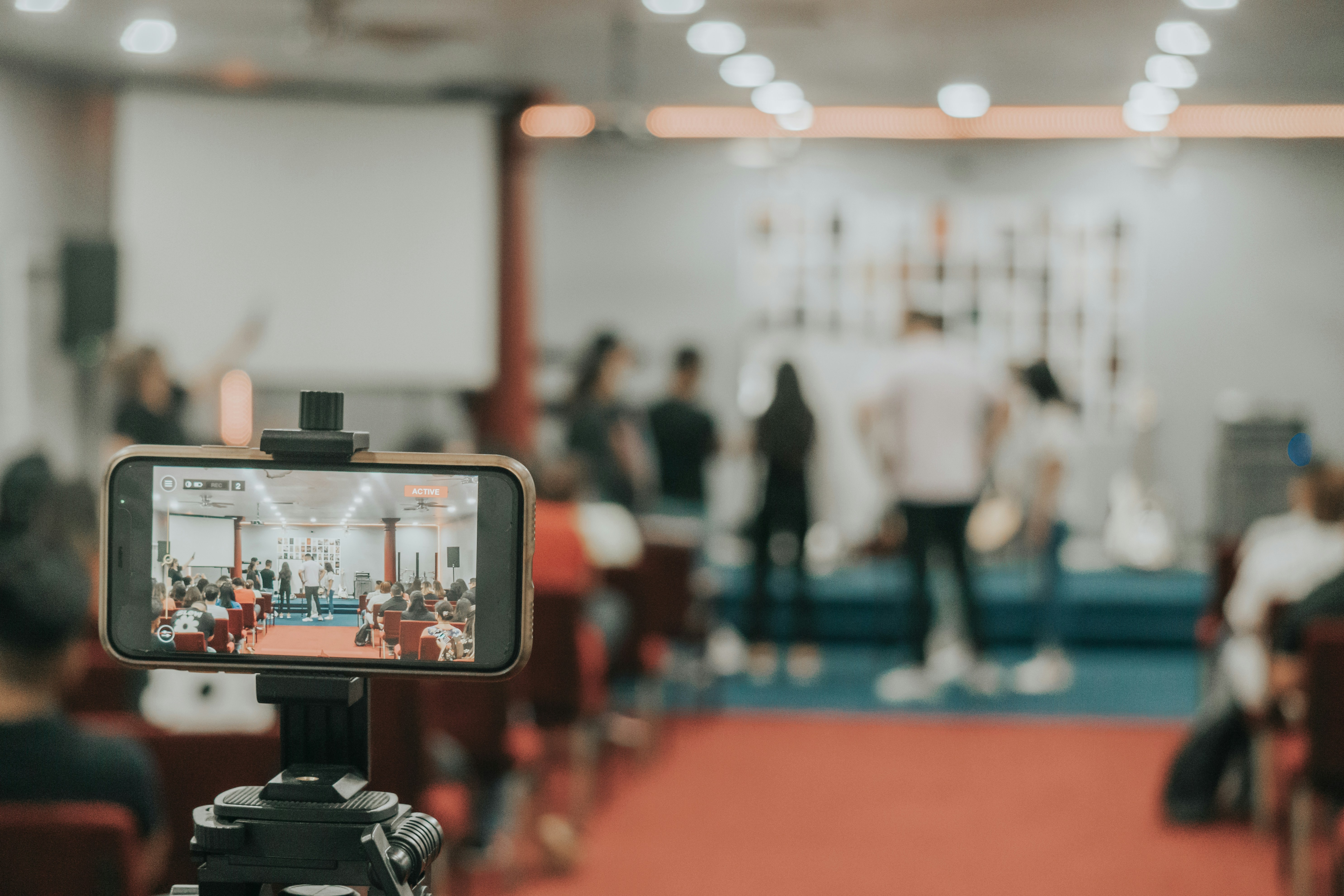 A smartphone mounted on a tripod actively recording a live talk, with the speaker and seated audience visible on screen